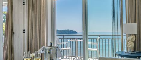 A balcony with a view of the sea, a table with a bottle, glasses, and a plate of food.