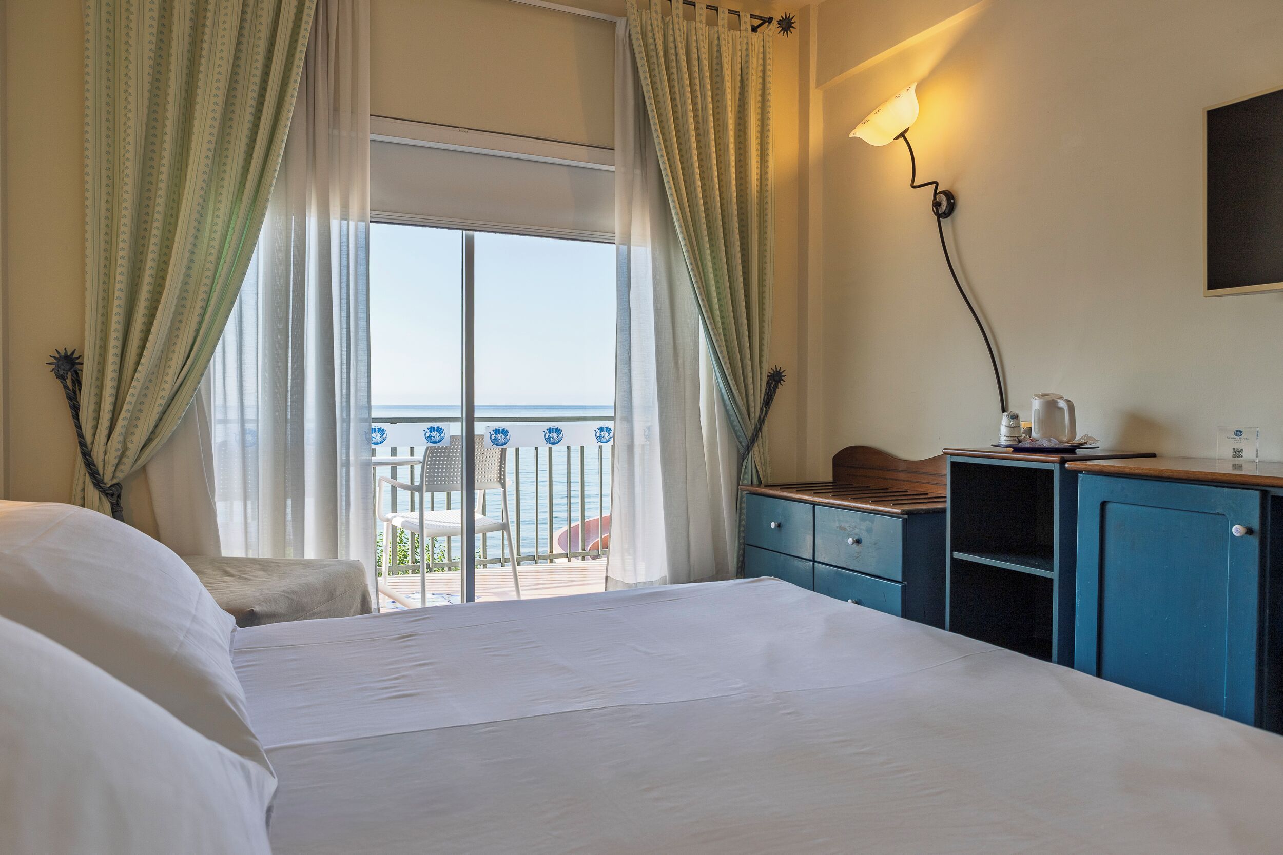 Habitación de hotel con cama, escritorio, silla y vista al mar a través de un gran ventanal.