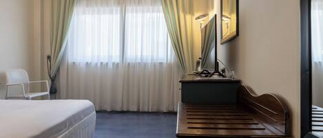 Habitación de hotel con cama, un banco de madera, un escritorio con espejo y dos ventanas con cortinas.