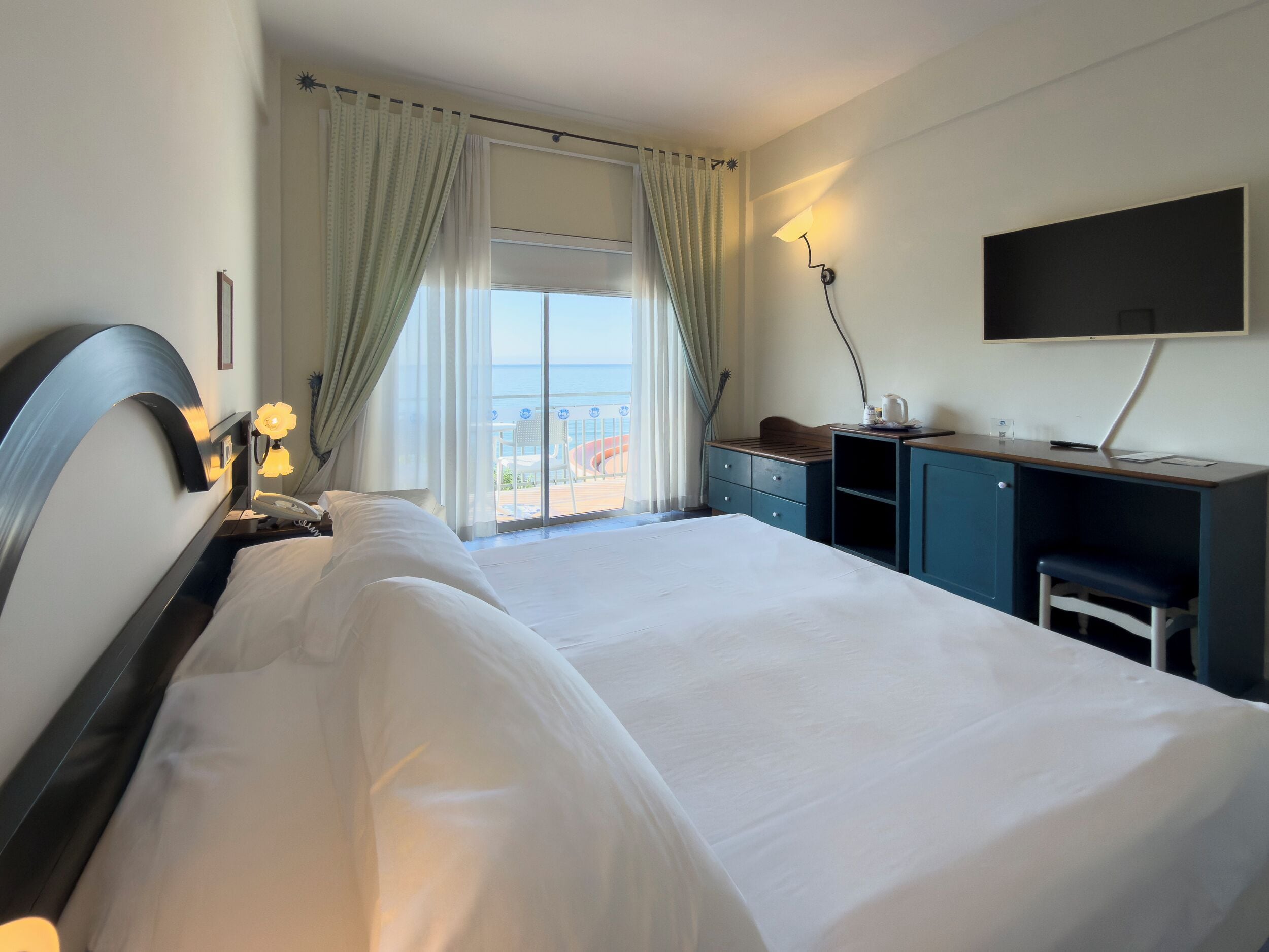 Habitación de hotel con cama, escritorio, silla, televisor y vista al mar a través de la ventana.