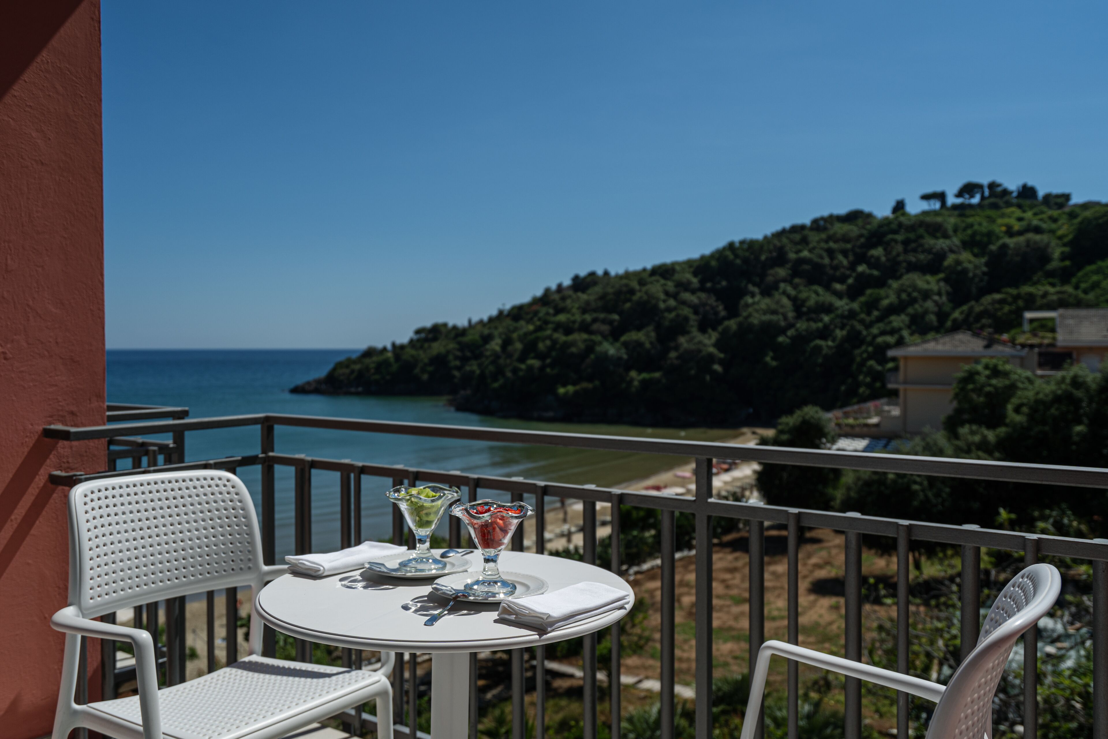 A balcony with a white table and chairs, overlooking a coastal landscape with trees and a clear sky.