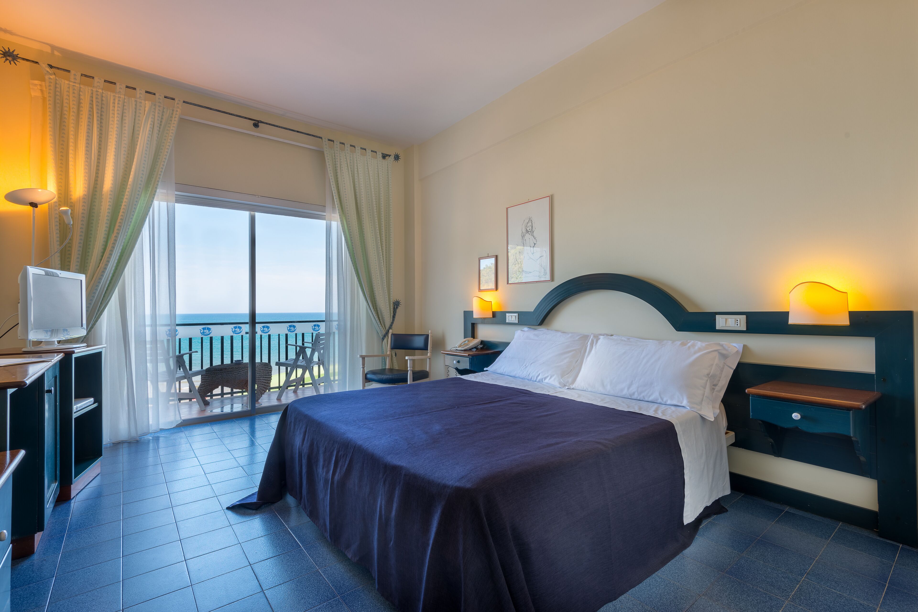 A hotel room with a bed, bedside table, lamp, and a TV. There is a balcony with a chair and a table.