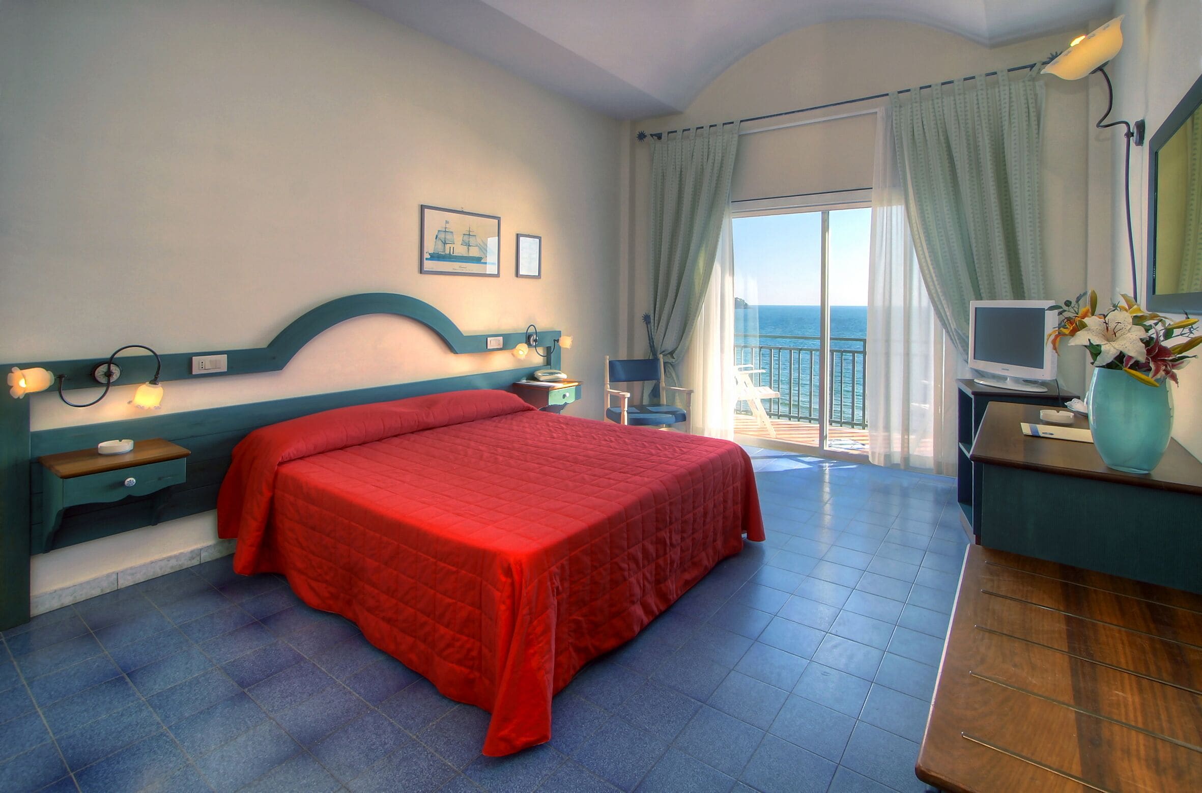 Ein Hotelzimmer mit Bett, Schreibtisch mit Computer, Stuhl, Fenster mit Vorhängen und Meerblick.