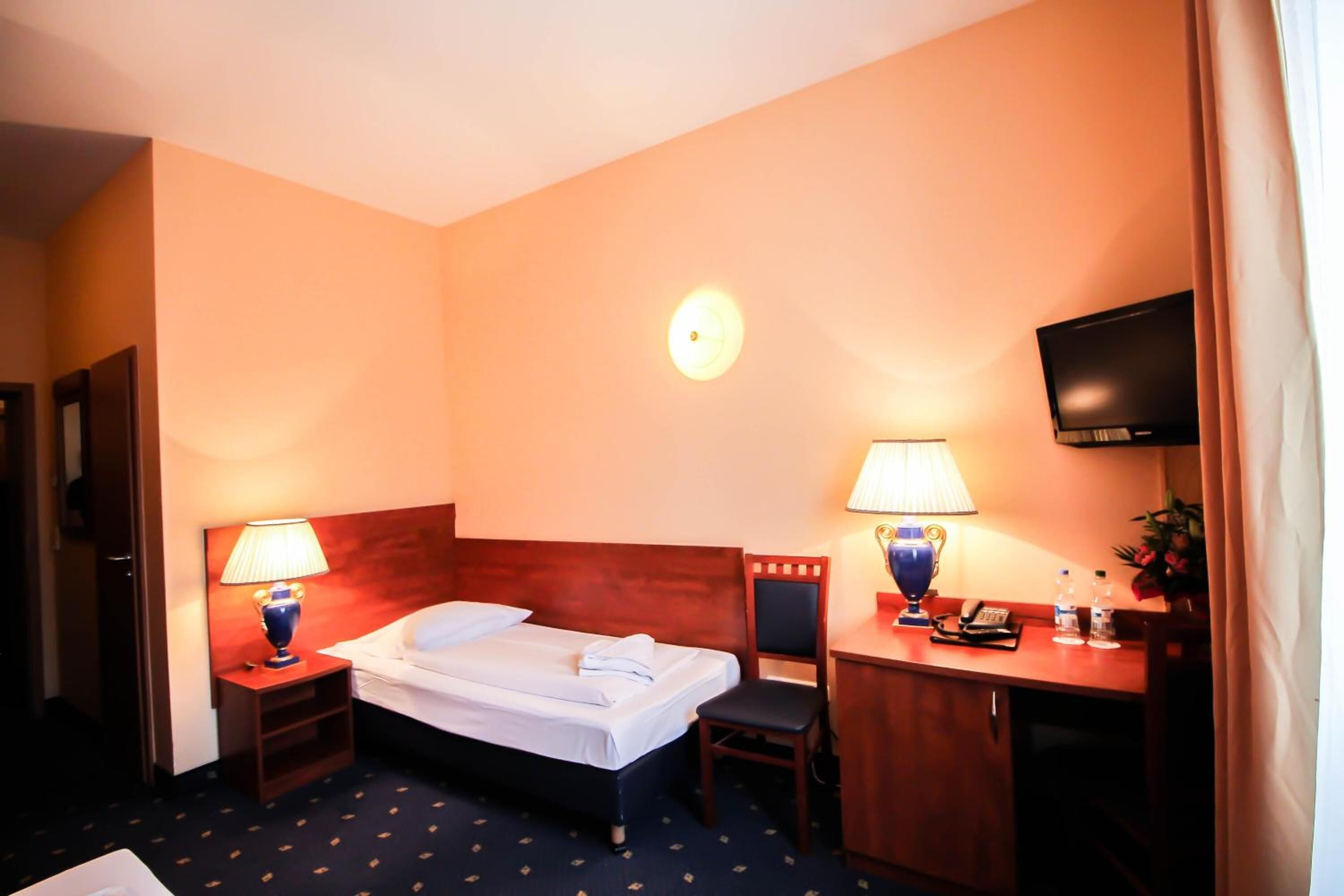 A hotel room with a bed, two bedside tables with lamps, a desk with a chair, a television, and a small cabinet.