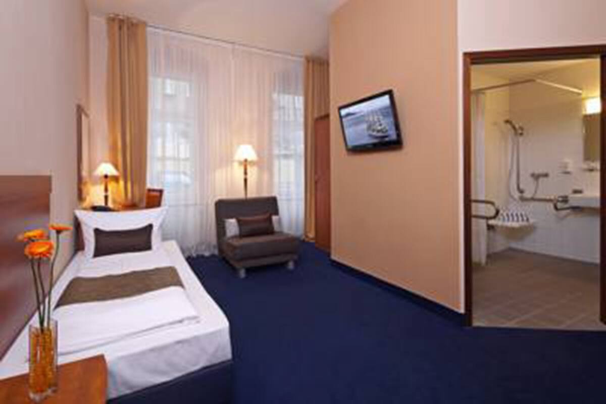 A hotel room with a bed, a chair, a desk with a vase of flowers, a television on the wall, and a bathroom visible through an open door.