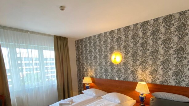 A hotel room with a double bed, two bedside lamps, a grey sofa, and a patterned wallpaper. - Hotel Atrium Charlottenburg (Berlin)