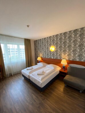 A hotel room with a double bed, two bedside lamps, a grey sofa, and a patterned wallpaper. - Hotel Atrium Charlottenburg (Berlin)