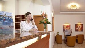 A hotel reception desk with a receptionist, a reception area with a wooden counter, a vase with flowers, and a wall with framed pictures. - Hotel Atrium Charlottenburg (Berlin)
