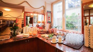 A buffet setup with various dishes and drinks, a large window with a view of greenery, and a nautical-themed decor including a rope and anchor. - Hotel Atrium Charlottenburg (Berlin)