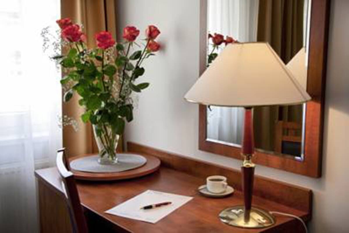 A hotel room with a wooden desk, a vase of red roses, a lamp, a cup of coffee, and a mirror.