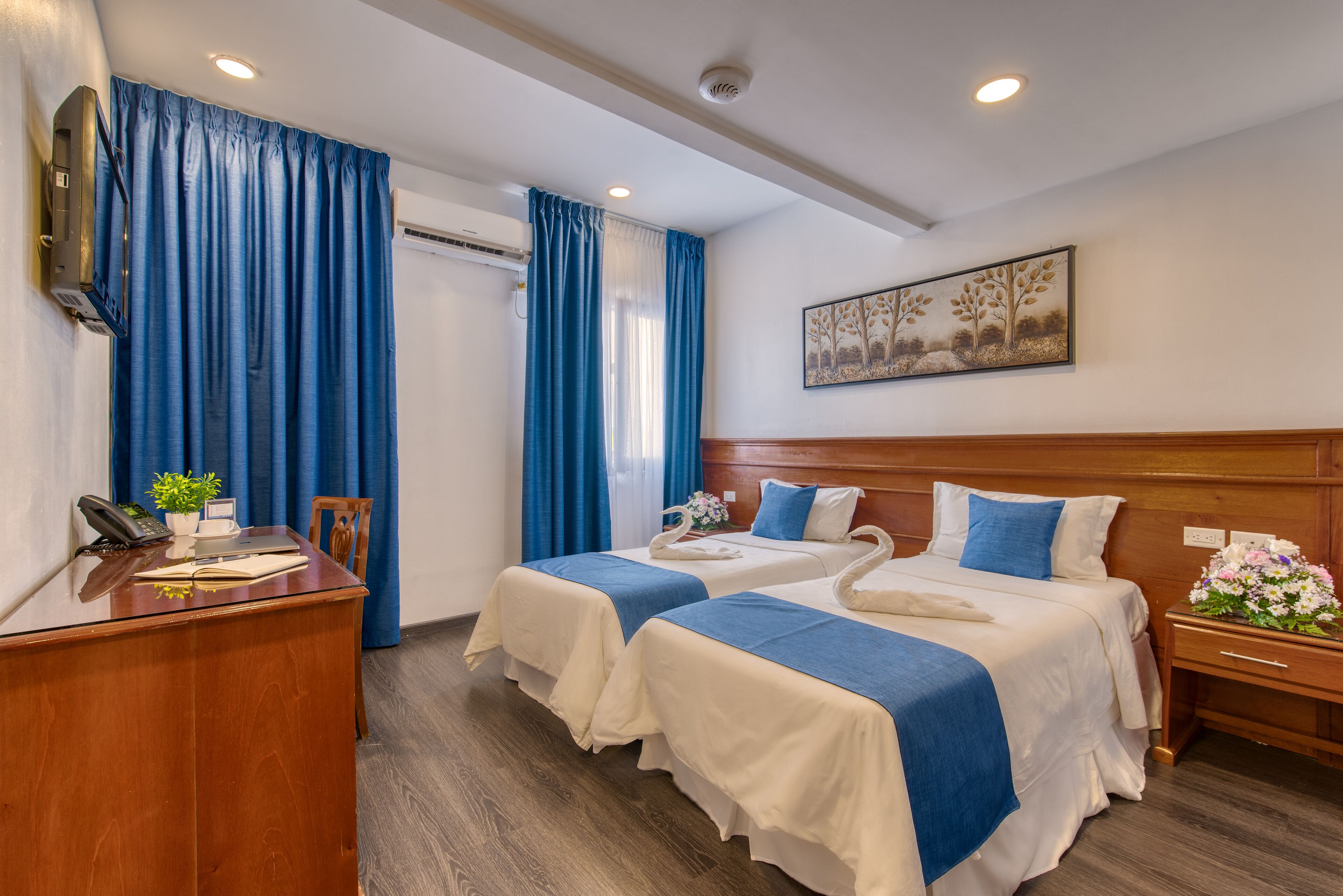 A hotel room with two beds, a desk, a telephone, a plant, and a painting on the wall.