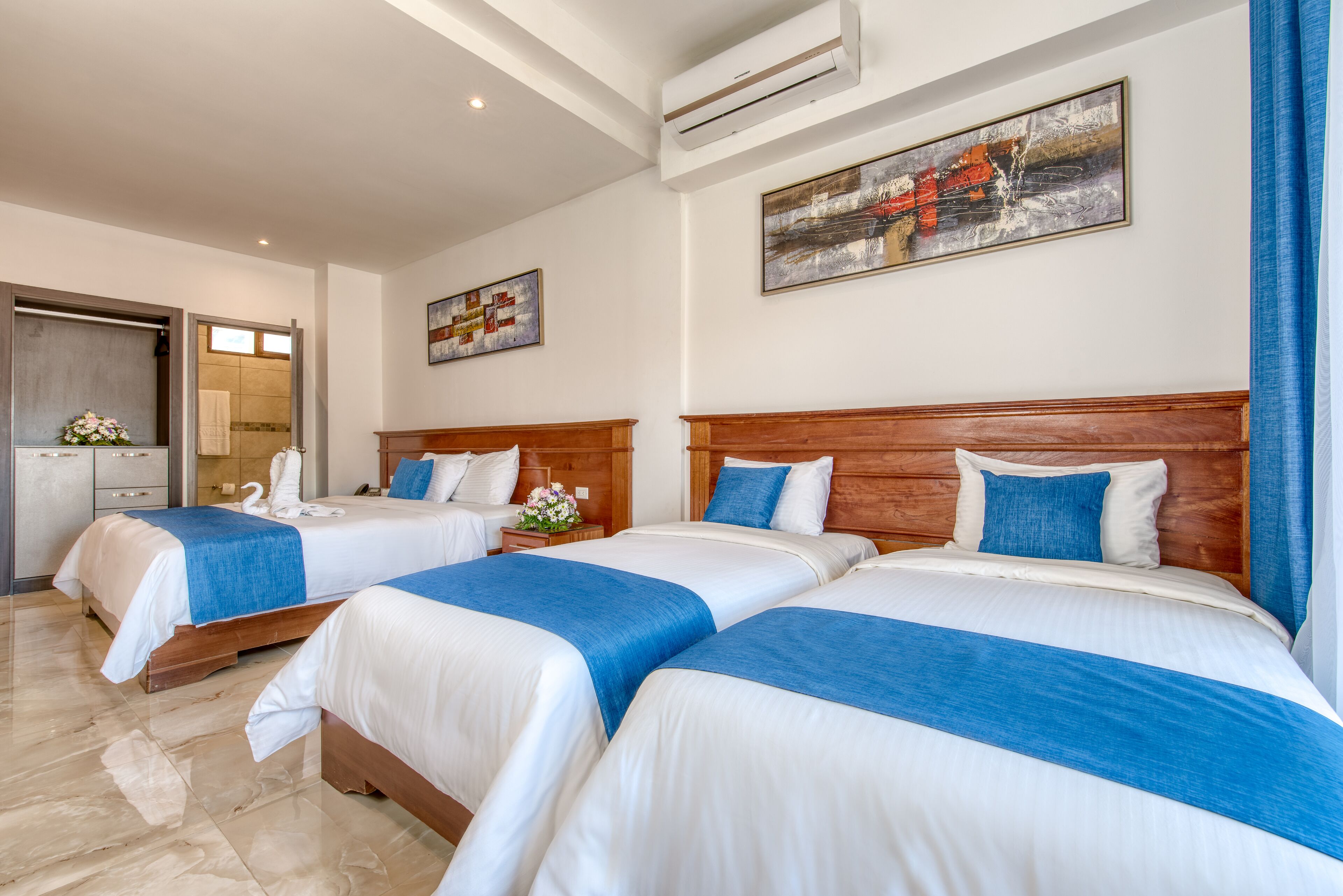Habitación de hotel con tres camas, cada una con cabecero de madera y ropa de cama azul y blanca. Hay un cuadro en la pared, un baño visible a través de una puerta abierta y una pequeña mesa con un florero.