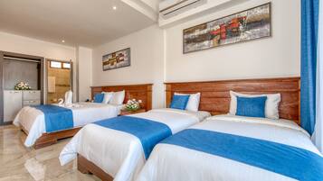 Camera d'albergo con tre letti, ognuno con testiera in legno e biancheria azzurra e bianca. Si nota un quadro appeso al muro, un bagno visibile attraverso una porta aperta e un tavolino con un vaso di fiori.