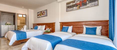 Habitación de hotel con tres camas, cada una con cabecero de madera y ropa de cama azul y blanca. Hay un cuadro en la pared, un baño visible a través de una puerta abierta y una pequeña mesa con un florero.