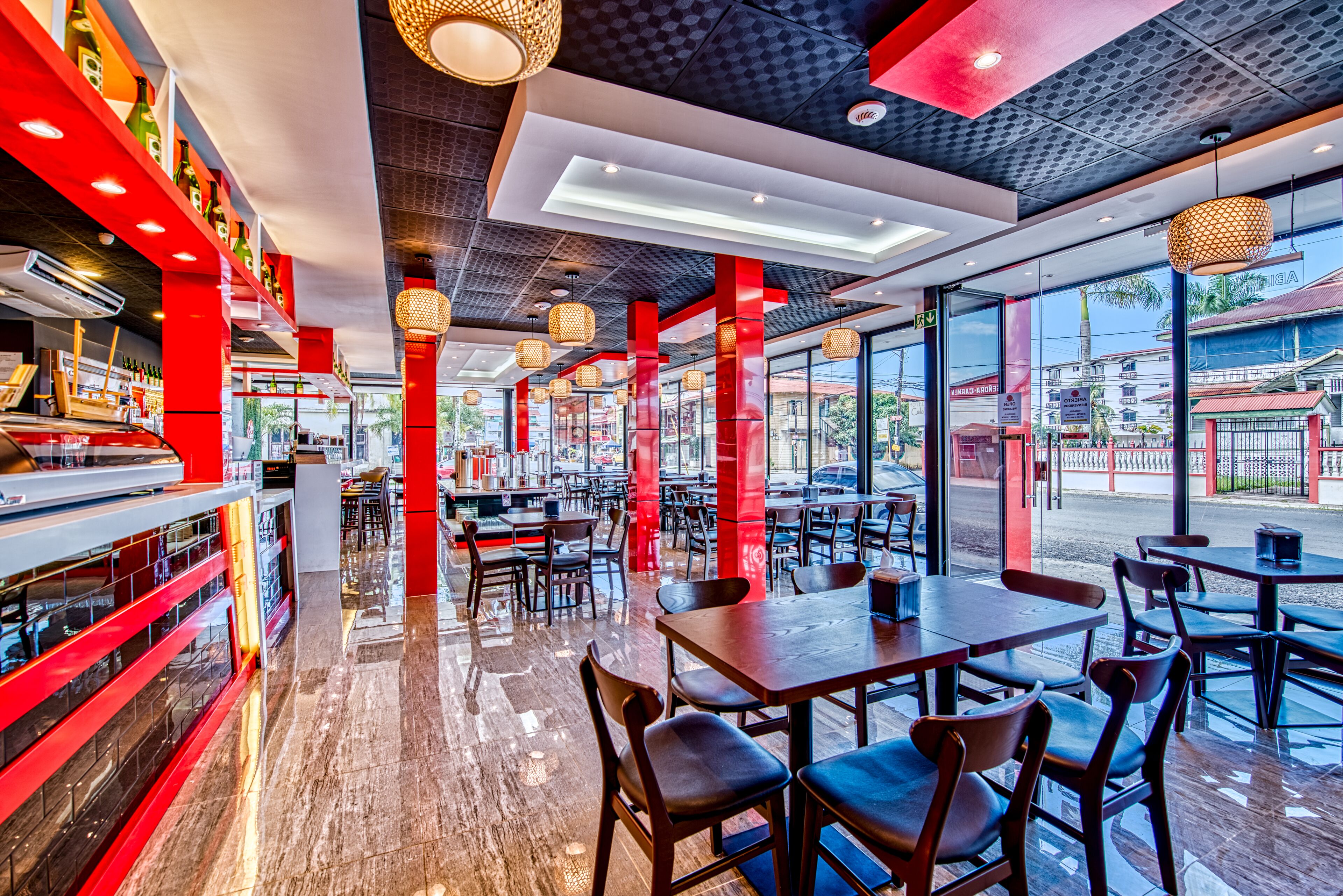 A modern restaurant interior with red accents, wooden tables and chairs, and large windows offering a view of the street.