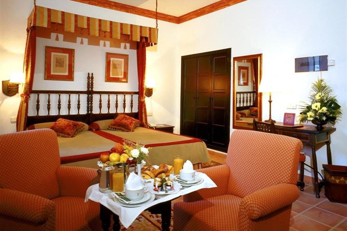 Ein Hotelzimmer mit Bett, zwei Sesseln, einem Esstisch mit Frühstück, einem Kleiderschrank und einem Spiegel.