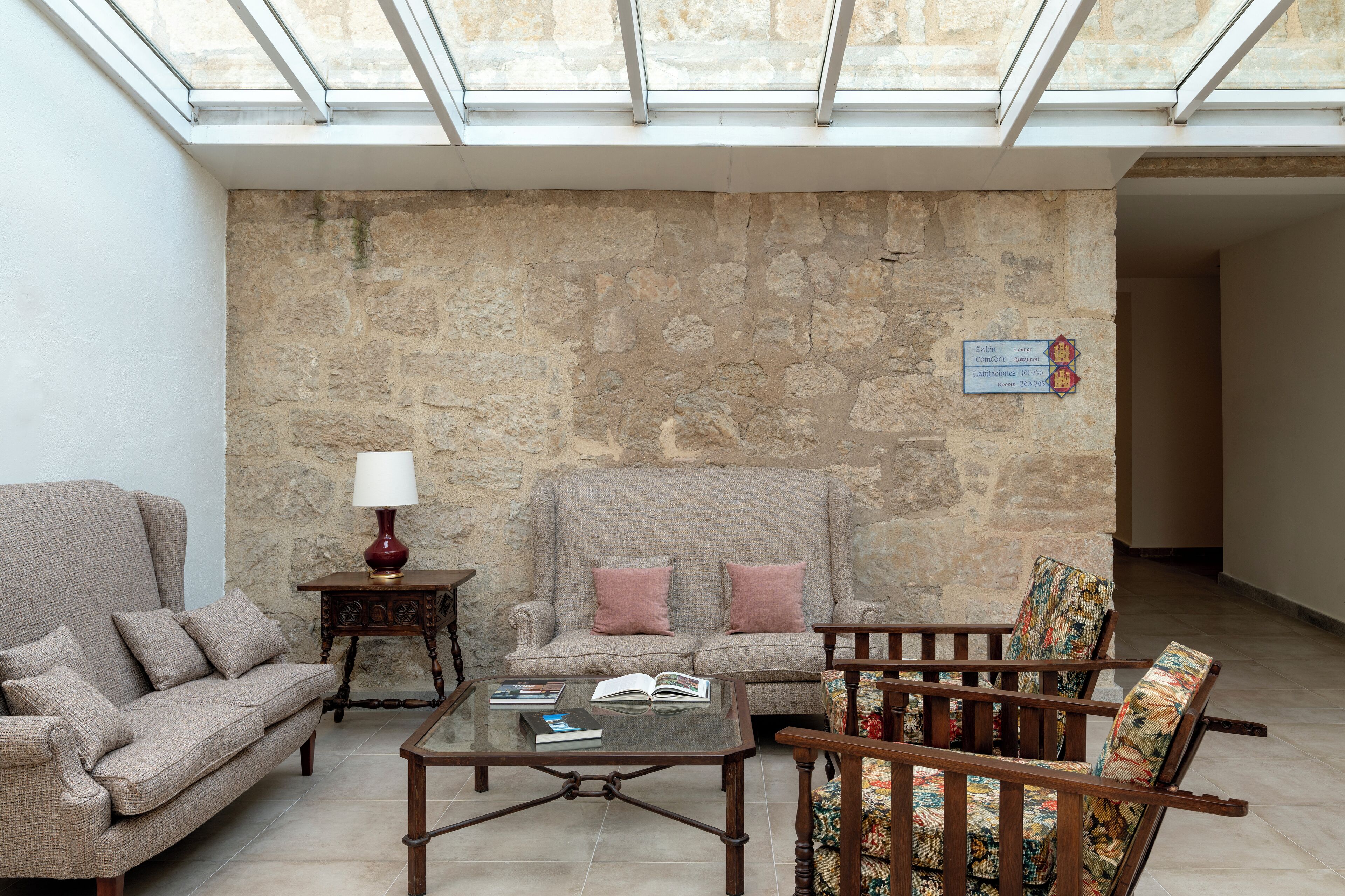 A room with a stone wall, a sofa, a chair, a small table with a lamp, and a book.