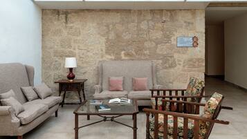 A room with a stone wall, a sofa, a chair, a small table with a lamp, and a book.