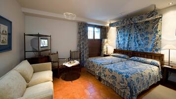 Habitación de hotel con sofá, dos camas con ropa de cama estampada, televisor sobre un mueble, mesa con bandeja y ventana con cortinas.