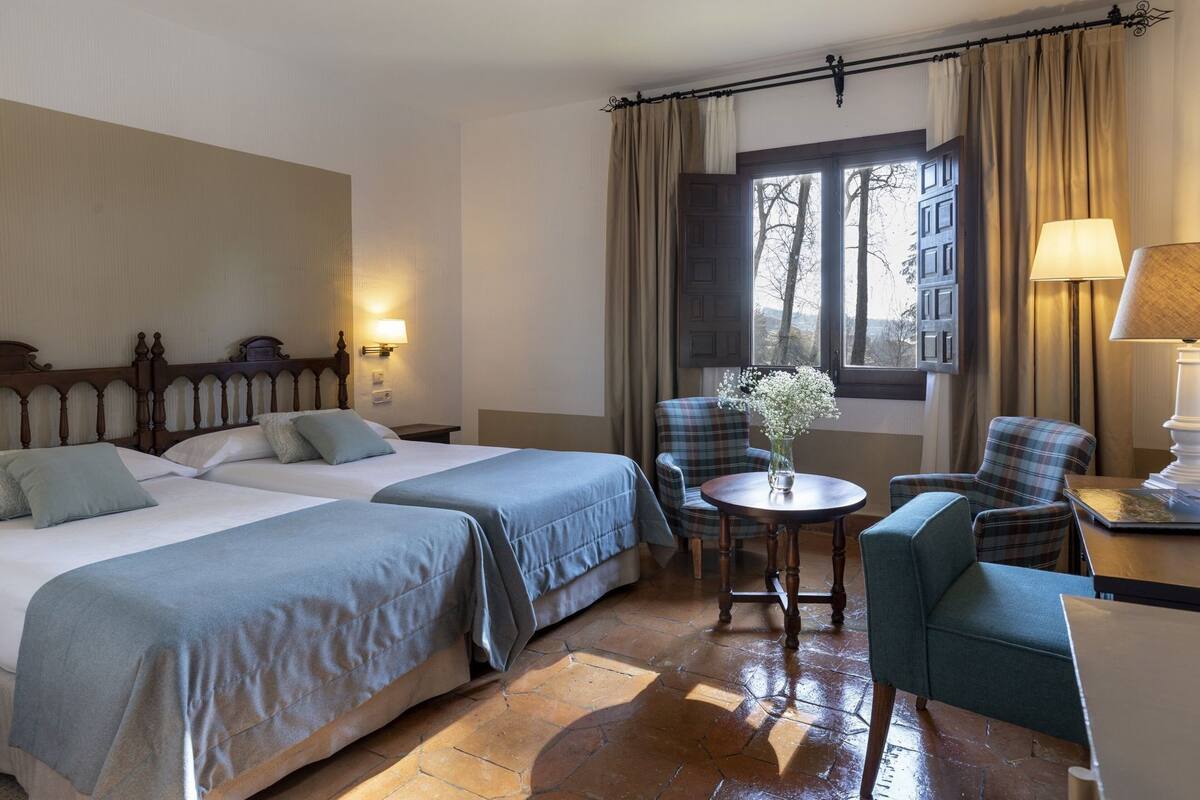 A hotel room with two beds, a round table, a chair, and a view of the outside.