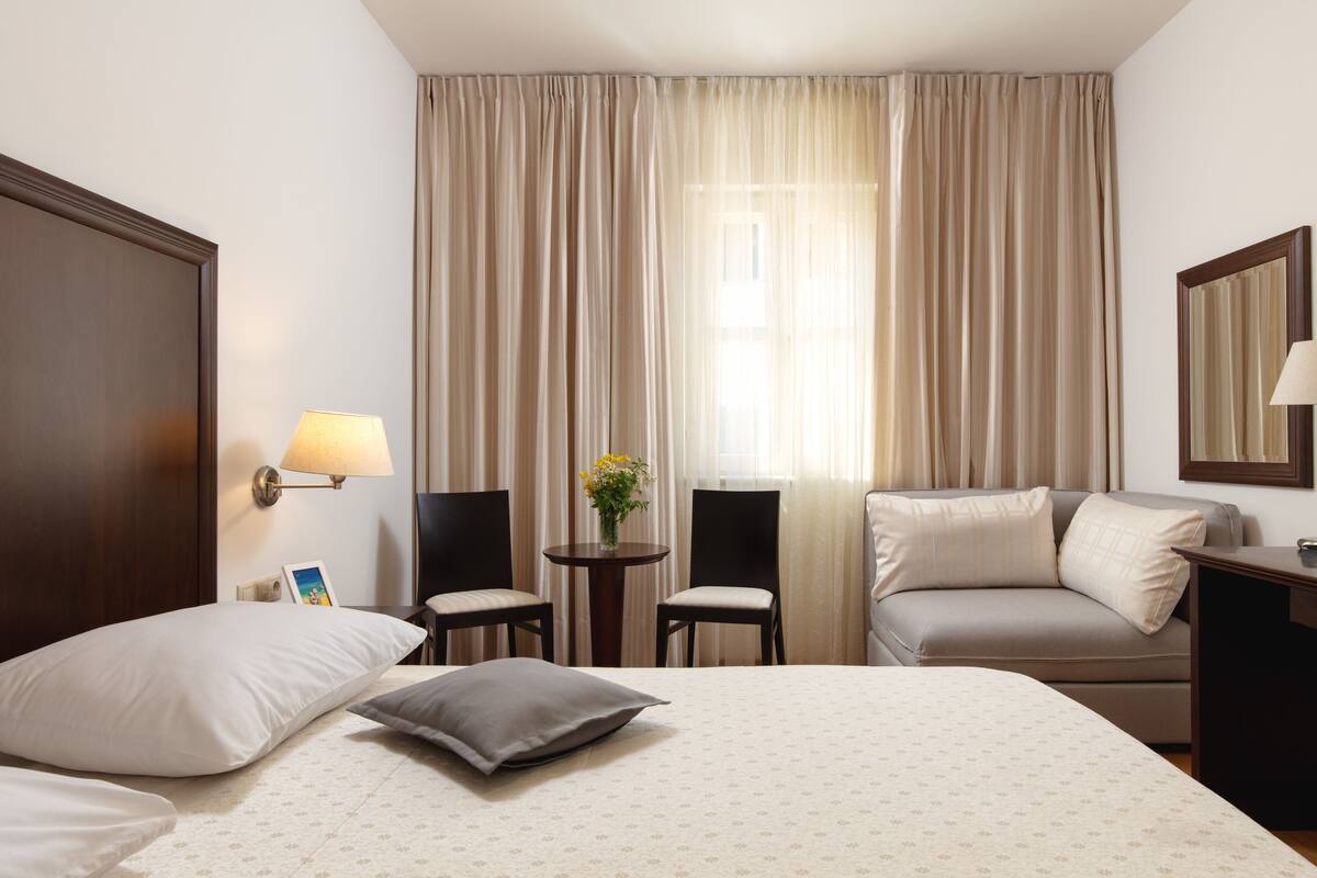A hotel room with a bed, two chairs, a small table with a vase, a sofa, a desk, and a lamp.