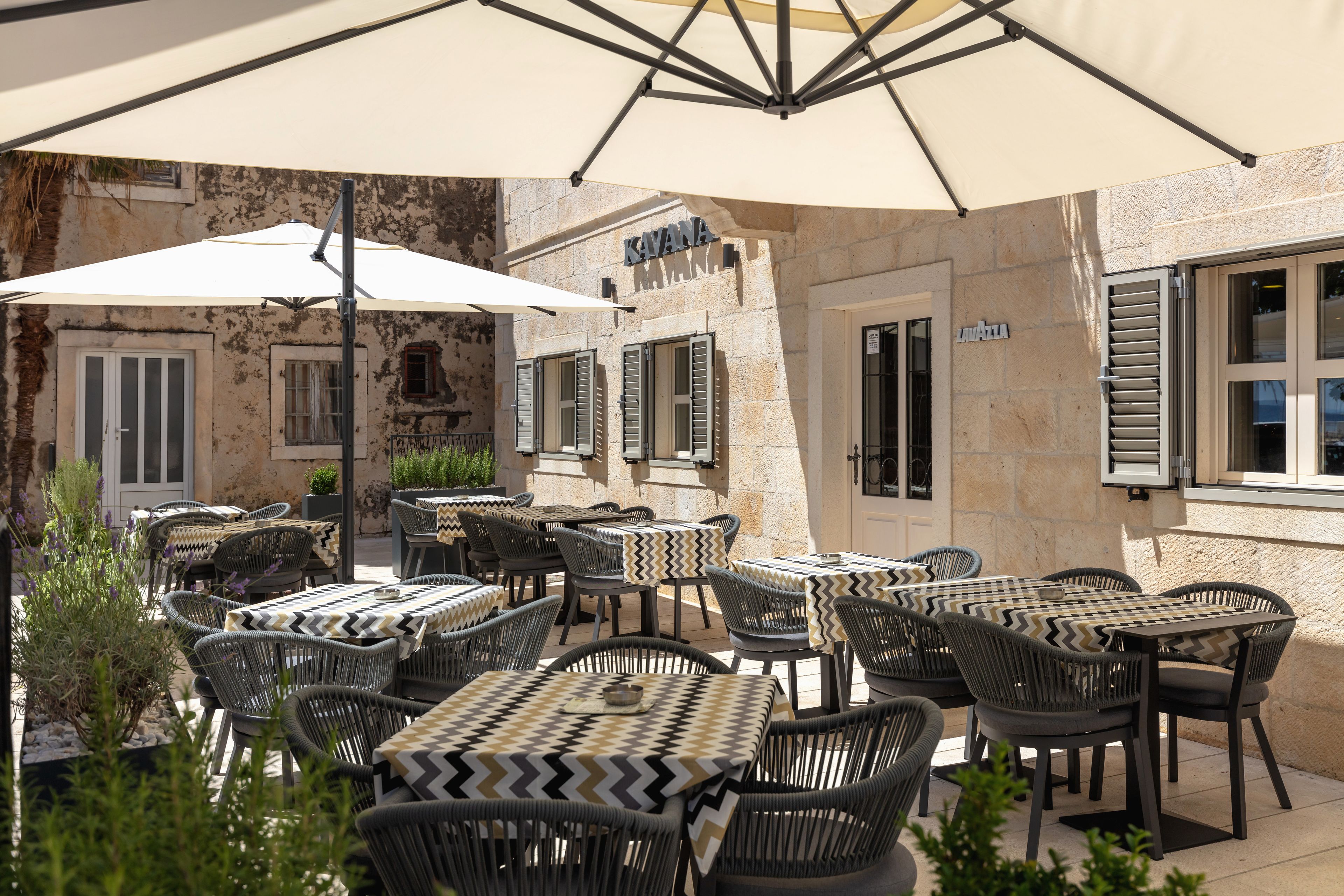 An outdoor seating area with tables and chairs, a large umbrella, and a building with a stone facade.
