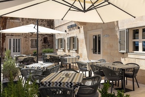 An outdoor seating area with tables and chairs, a large umbrella, and a building with a stone facade. - Hotel Croatia (Baska Voda)