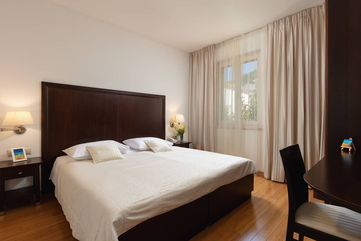 A neatly made bed with white linens, a wooden headboard, two bedside lamps, a nightstand with a lamp, a small table with a book, and a chair.