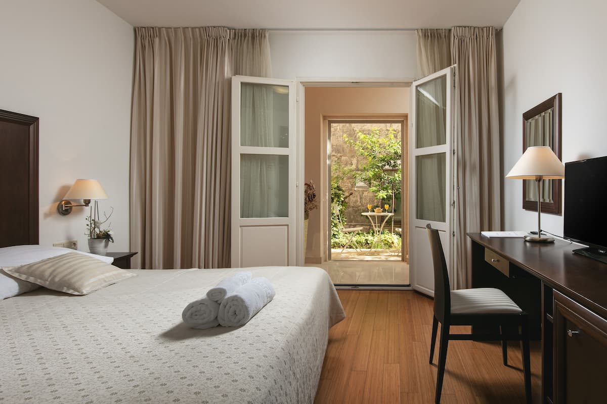 A hotel room with a bed, a desk, a chair, a TV, and a view of the garden through the open door.
