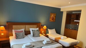 A hotel room with two beds, a wooden headboard, bedside lamps, a small painting, and a kitchenette. - 121 Paris Hotel (Paris)