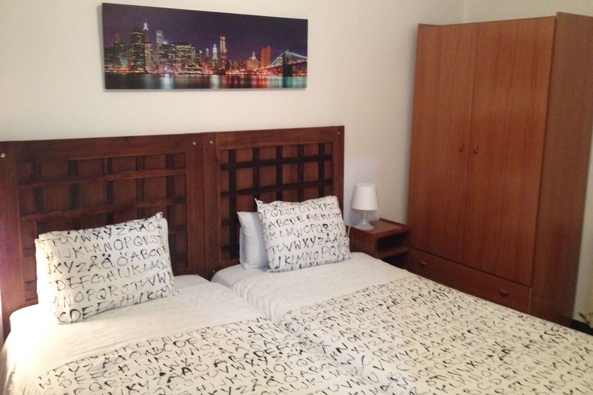 A bedroom with a cityscape painting, a wooden headboard, two pillows with alphabet patterns, a bedside table with a lamp, and a wooden wardrobe.