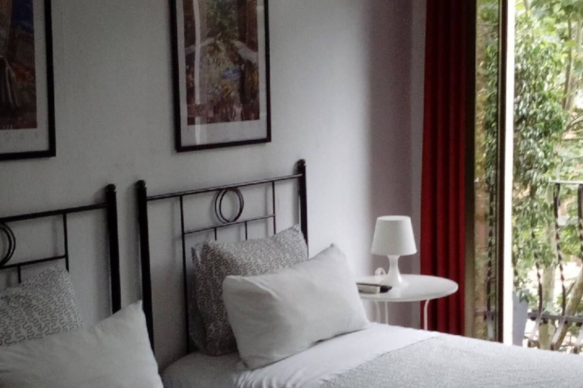 A hotel room with two beds, a small table, a lamp, and a window with curtains.