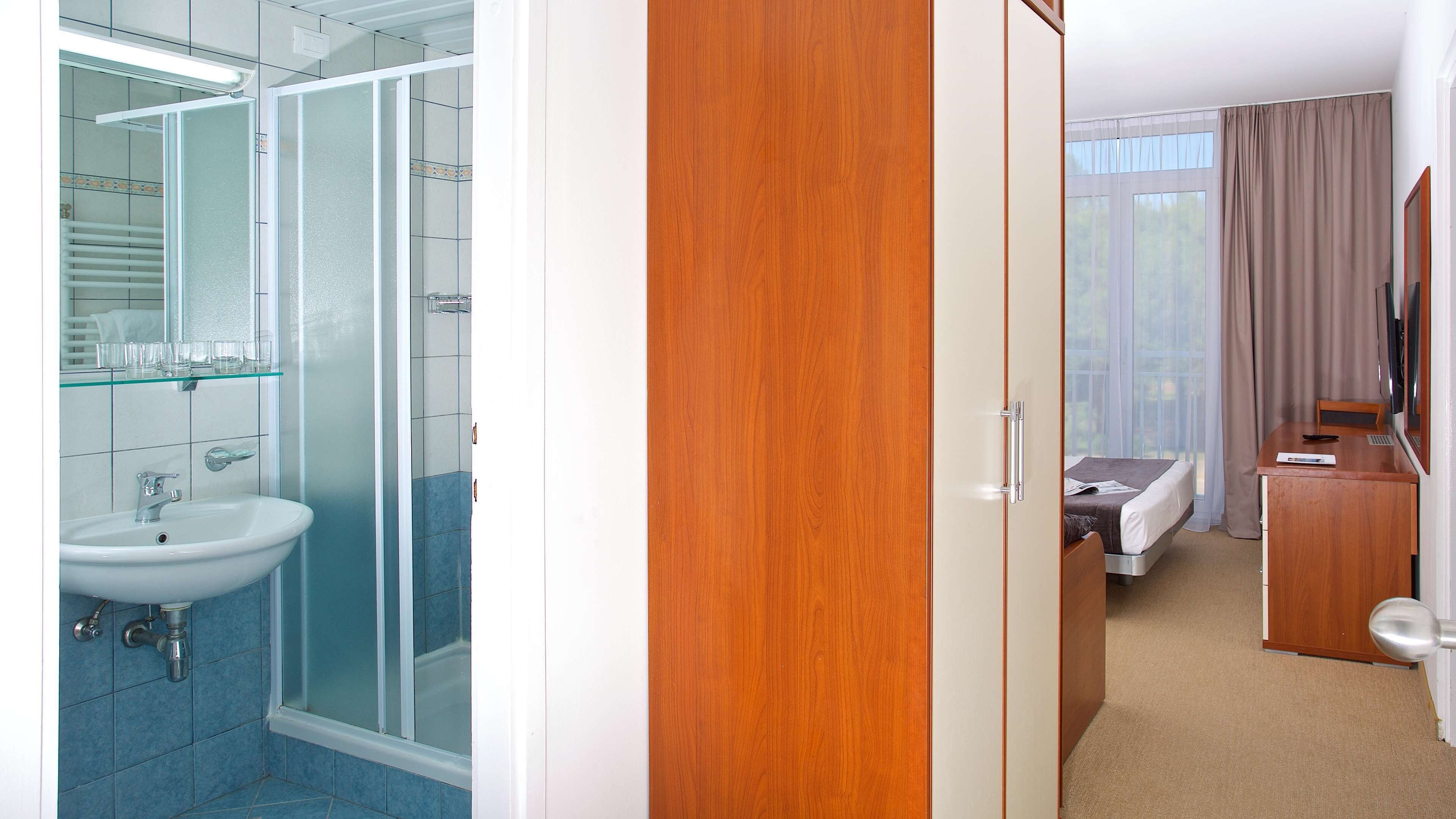 A hotel room with a bathroom, a wooden wardrobe, a bed, and a desk with a chair.