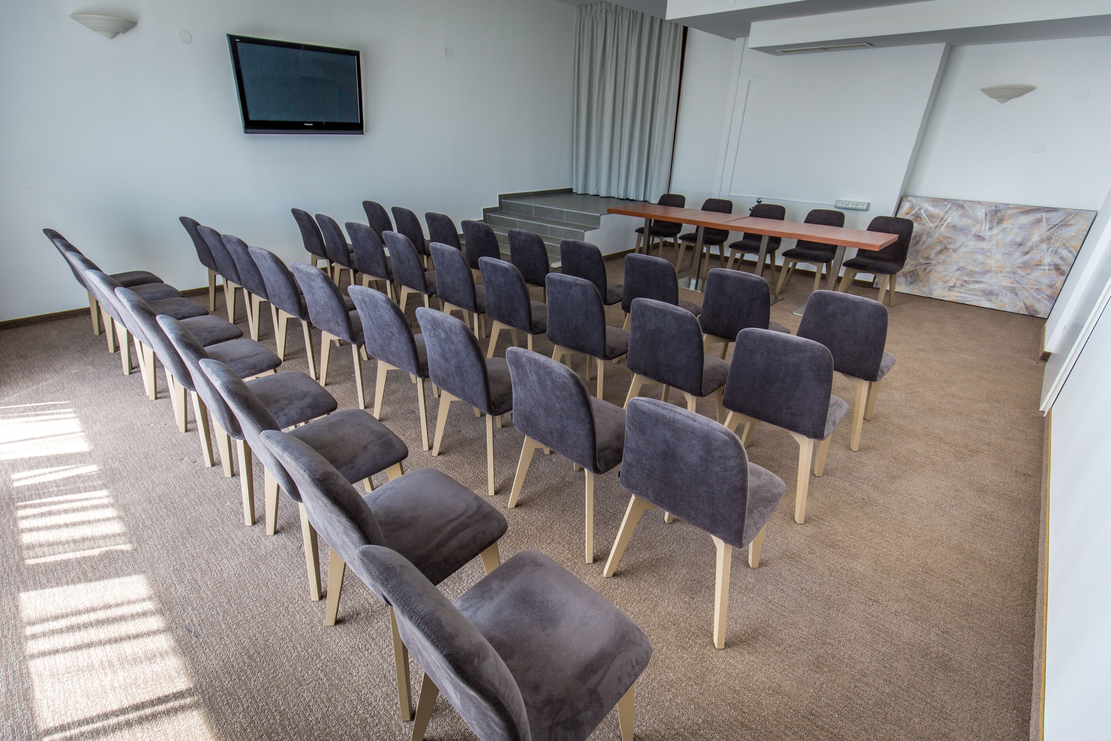 A conference room with rows of chairs, a long table, a projector screen, and a large window with curtains.