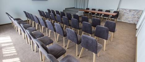 A conference room with rows of chairs, a long table, a projector screen, and a large window with curtains.