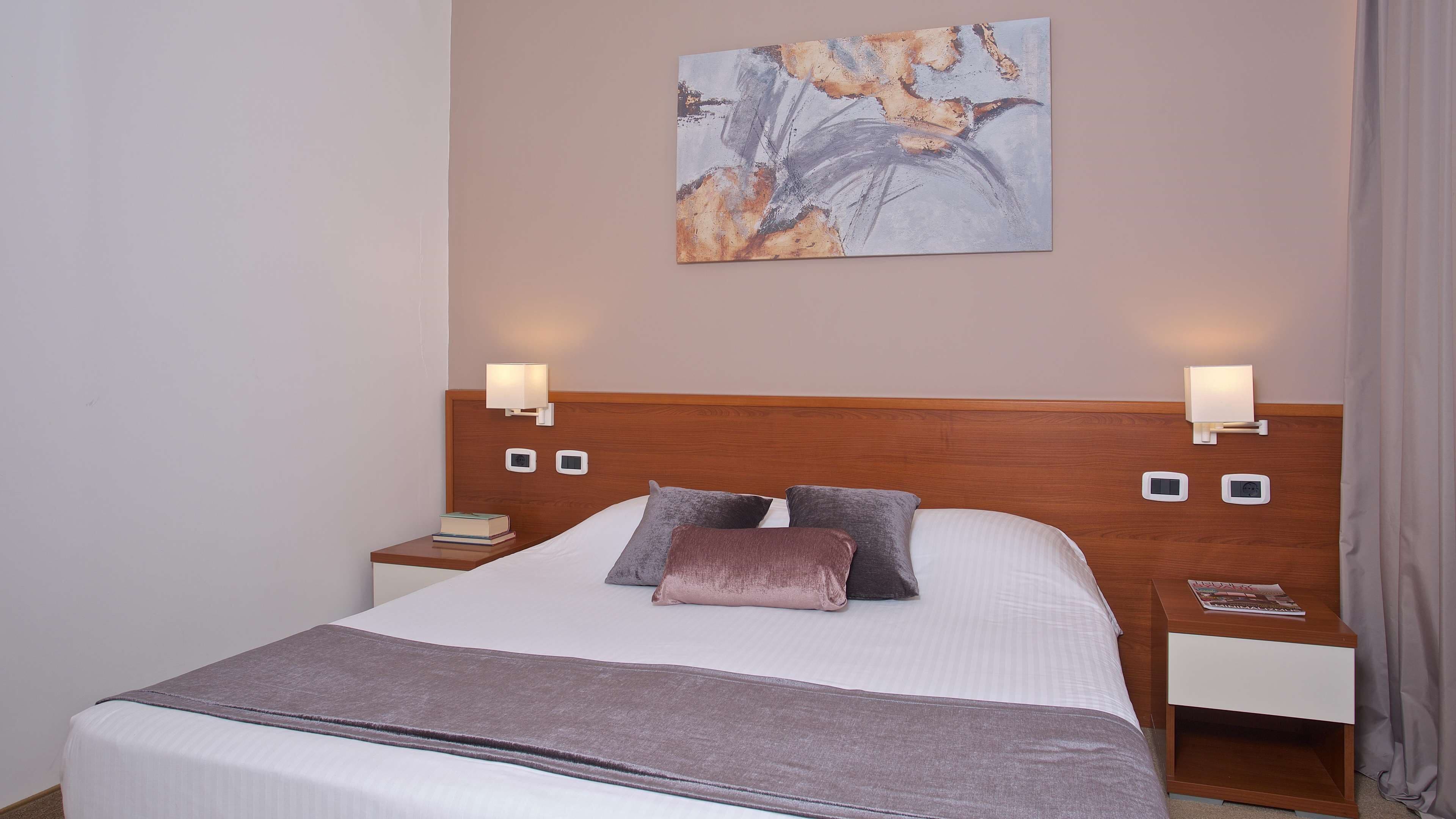 A neatly made bed with a wooden headboard, two bedside tables with lamps, a single artwork on the wall, and a window with curtains.