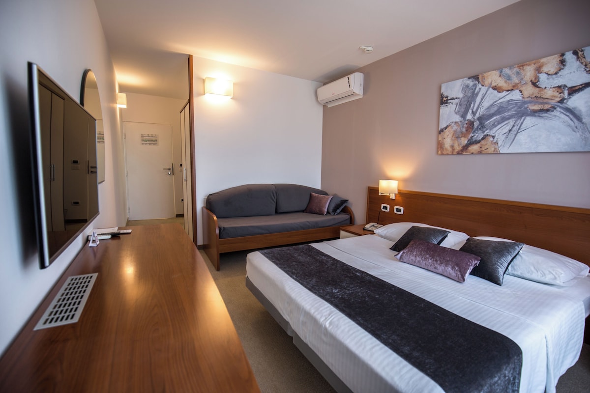 A modern hotel room with a large bed, a sofa bed, a flat-screen TV, and a painting on the wall.
