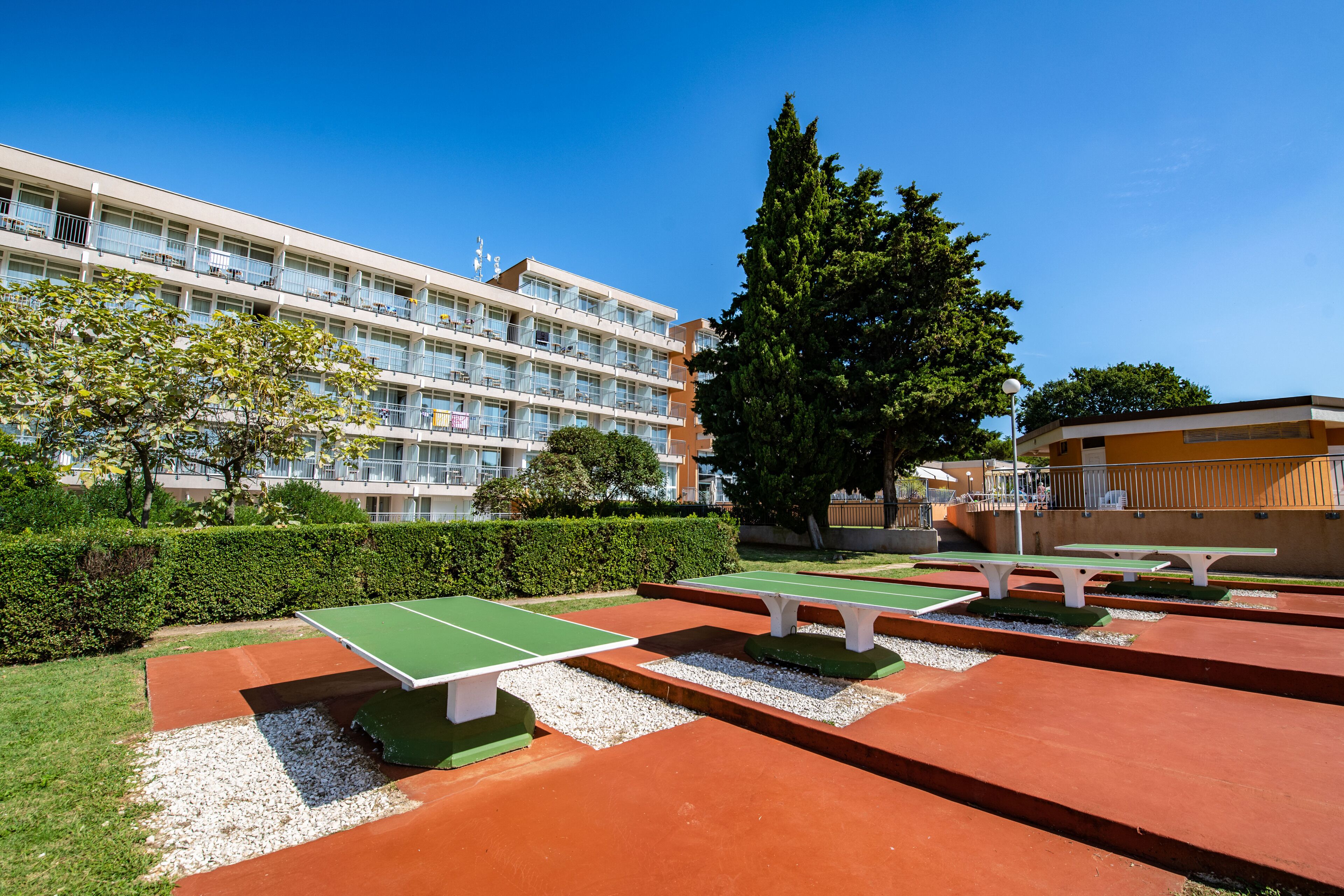 Un área recreativa al aire libre con mesas de ping pong y bancos, rodeada por un seto y un edificio con varios balcones.