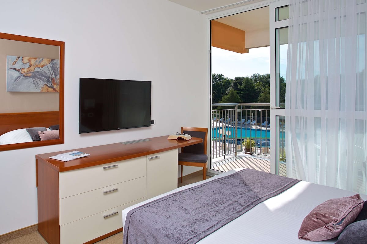 Une chambre d’hôtel équipée d’un lit, d’un bureau, d’une télévision, d’un miroir et d’un balcon donnant sur la piscine.