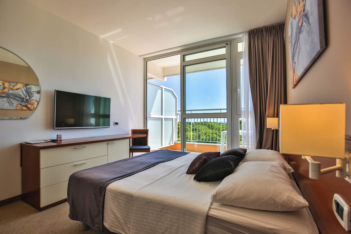Une chambre d’hôtel avec un lit, un bureau, une télévision, un balcon avec vue et un tableau au mur.