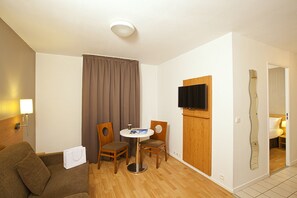A hotel room with a wooden floor, a brown sofa, a small round table with two chairs, a TV mounted on the wall, and a door leading to a bathroom. - Séjours & Affaires Paris Ivry (Ivry-sur-Seine)