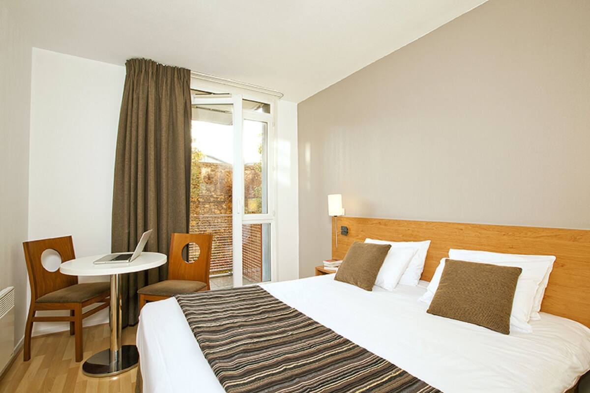 A hotel room with a bed, a desk with a laptop, a chair, and a window with curtains.