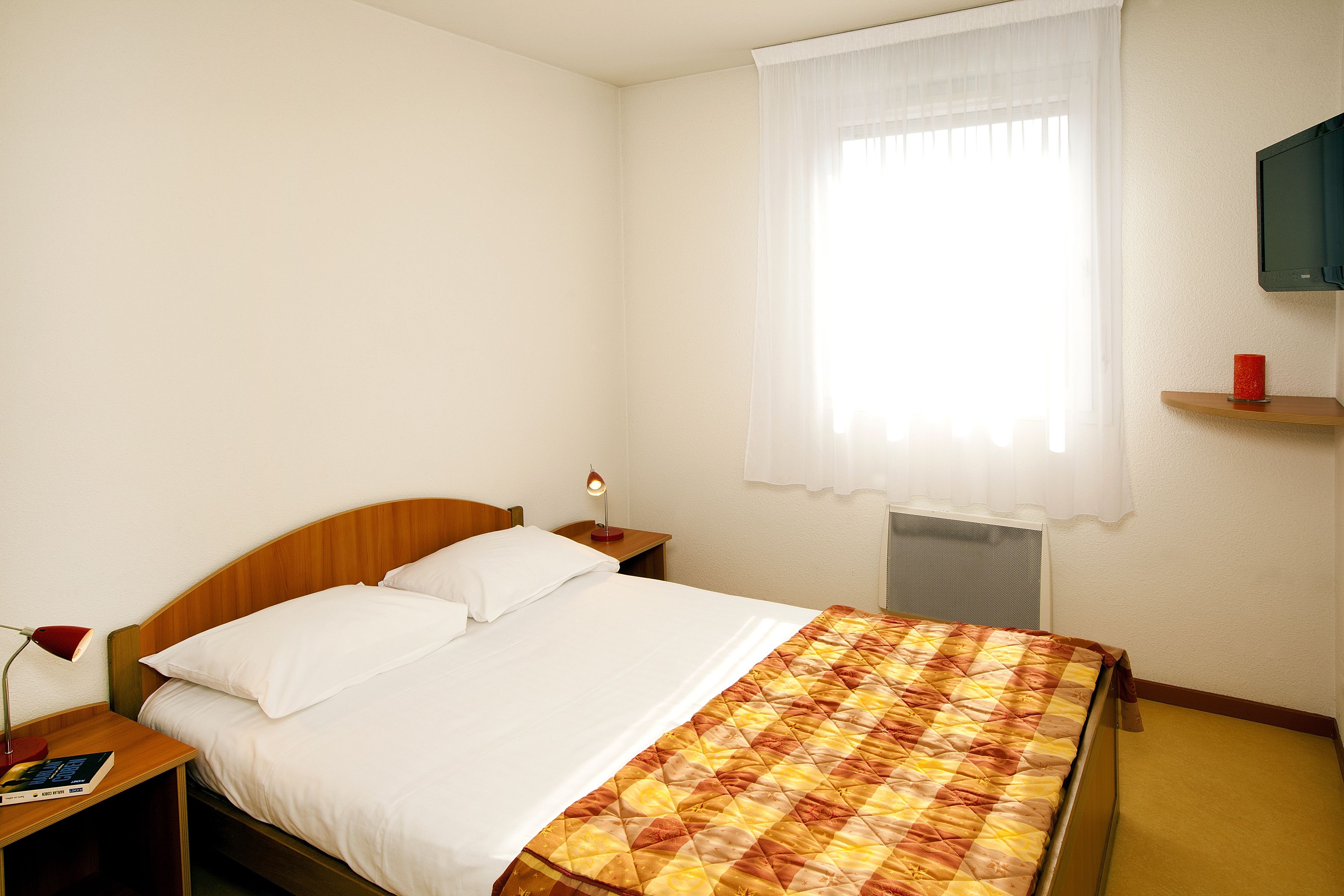 Habitación de hotel con cama, mesita de noche, lámpara, televisión en la pared y ventana con cortina.