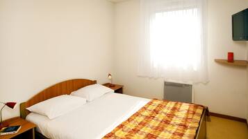 Habitación de hotel con cama, mesita de noche, lámpara, televisión en la pared y ventana con cortina.