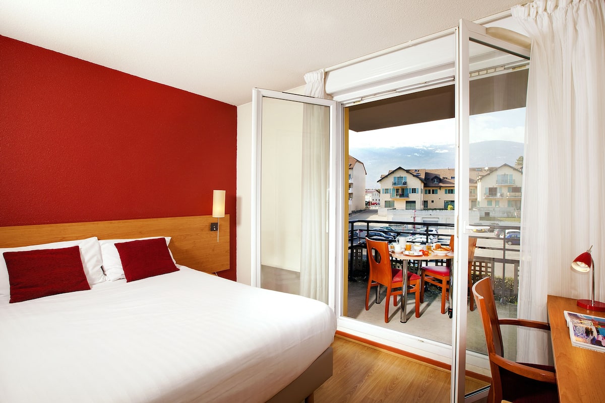 Ein Hotelzimmer mit einem Bett, einem Holzkopfteil, einem Balkon mit Tisch und Stühlen sowie Blick auf Gebäude.
