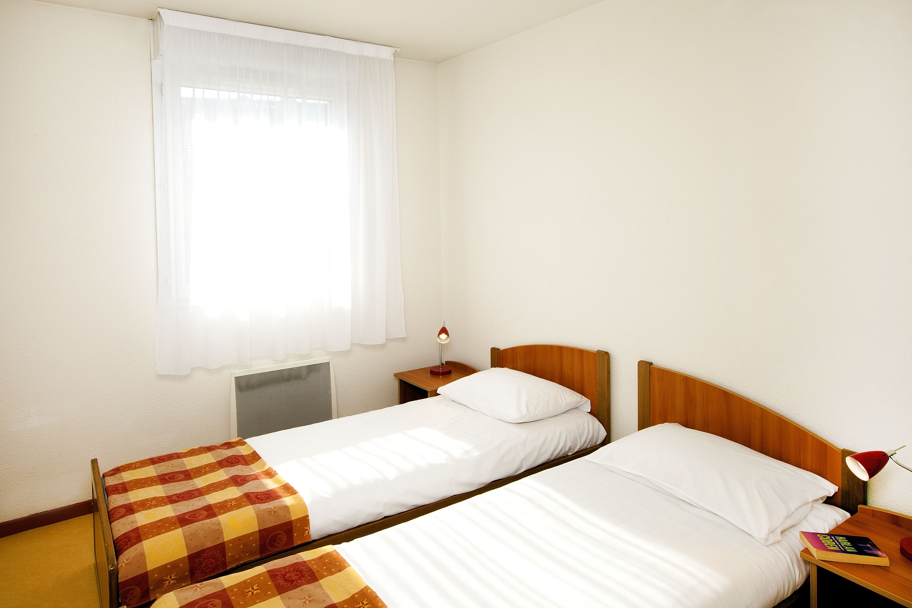Un letto matrimoniale con coperta a quadri e copripiumino bianco, un comodino con lampada e una finestra con tende trasparenti.