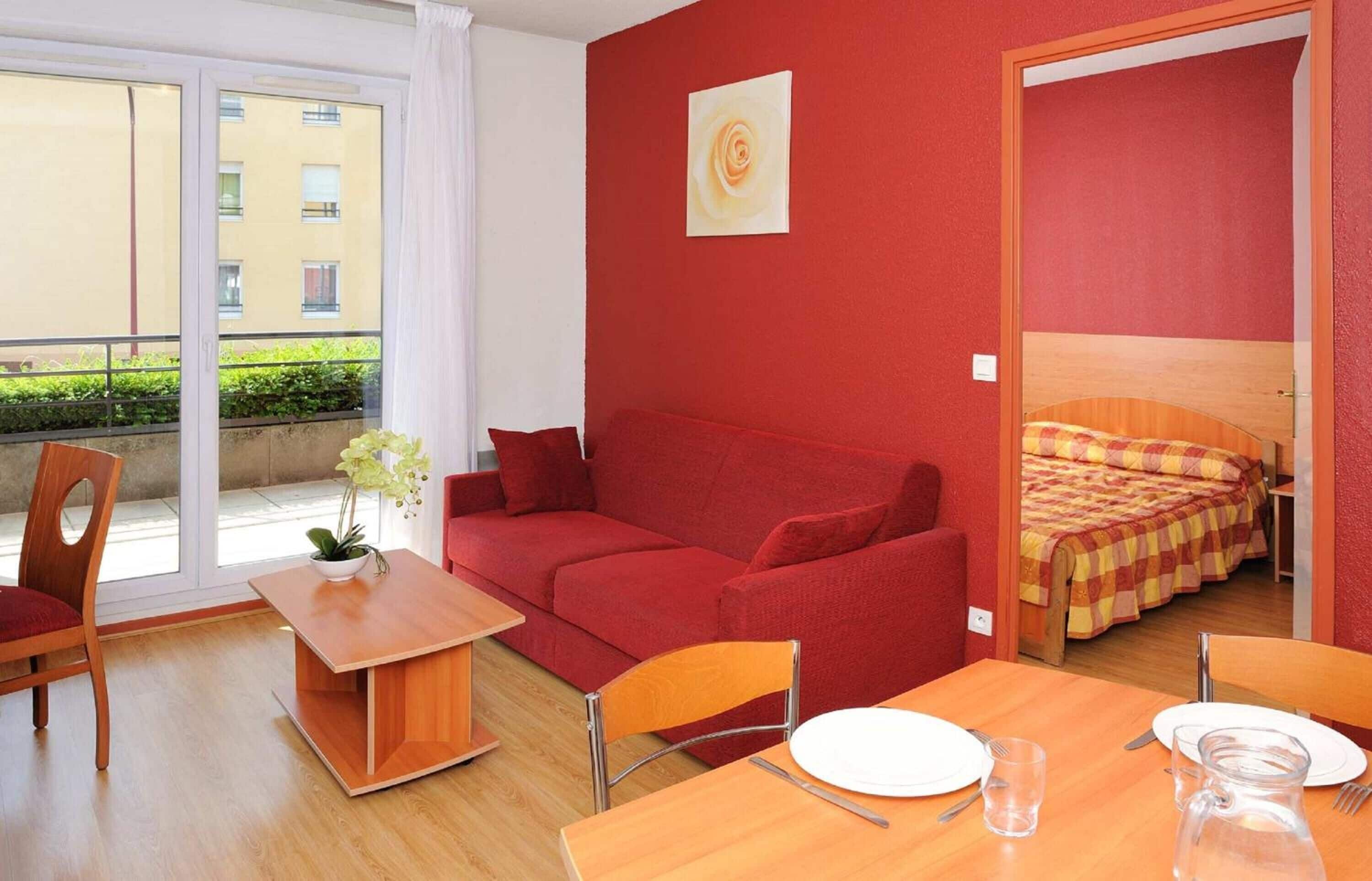 Una habitación pequeña con un sofá rojo, una mesa de madera con un jarrón de flores y una cama con una manta a cuadros.