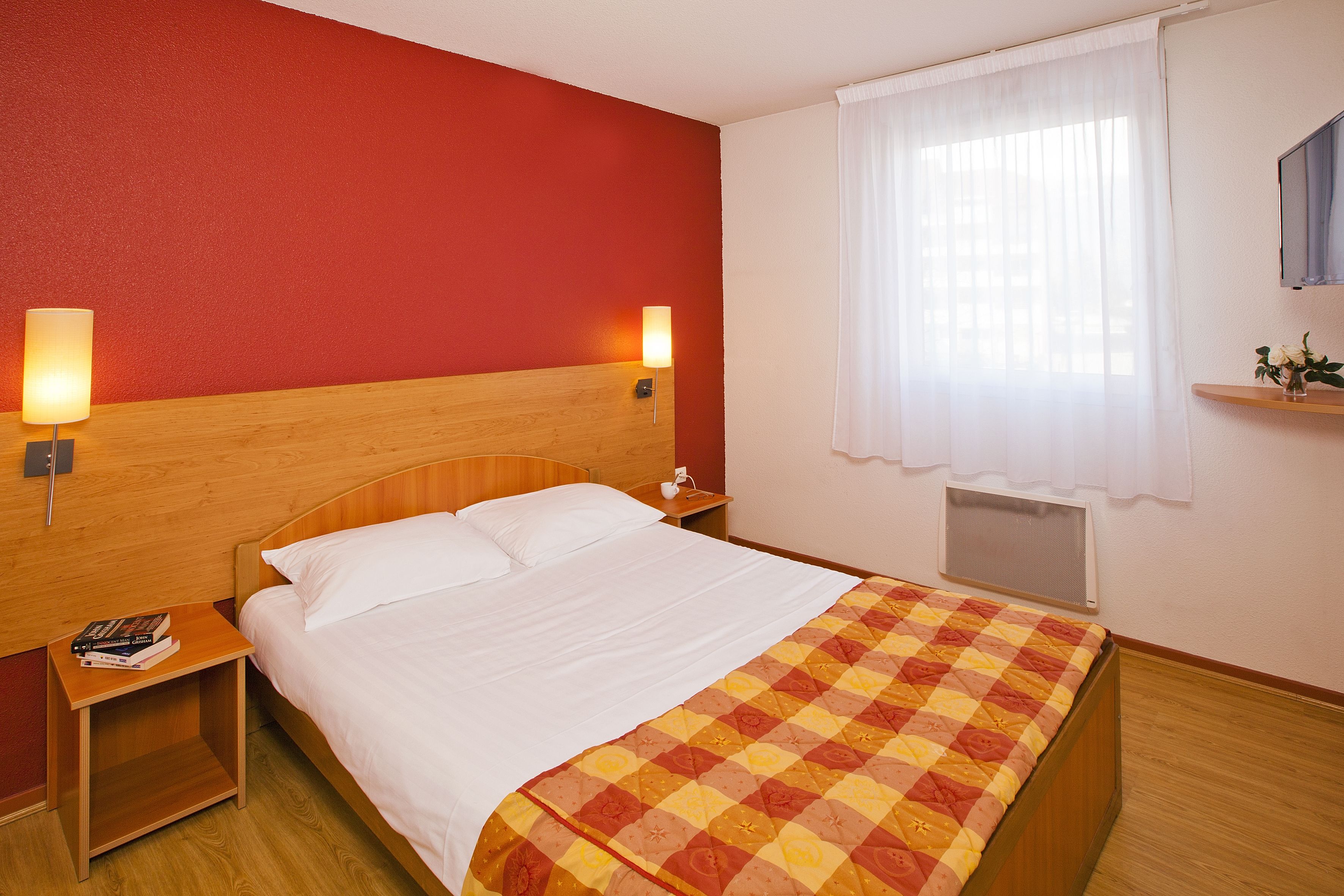 Habitación de hotel con cabecera de madera, colcha de cuadros, mesita de noche con lámpara y ventana con cortinas blancas.