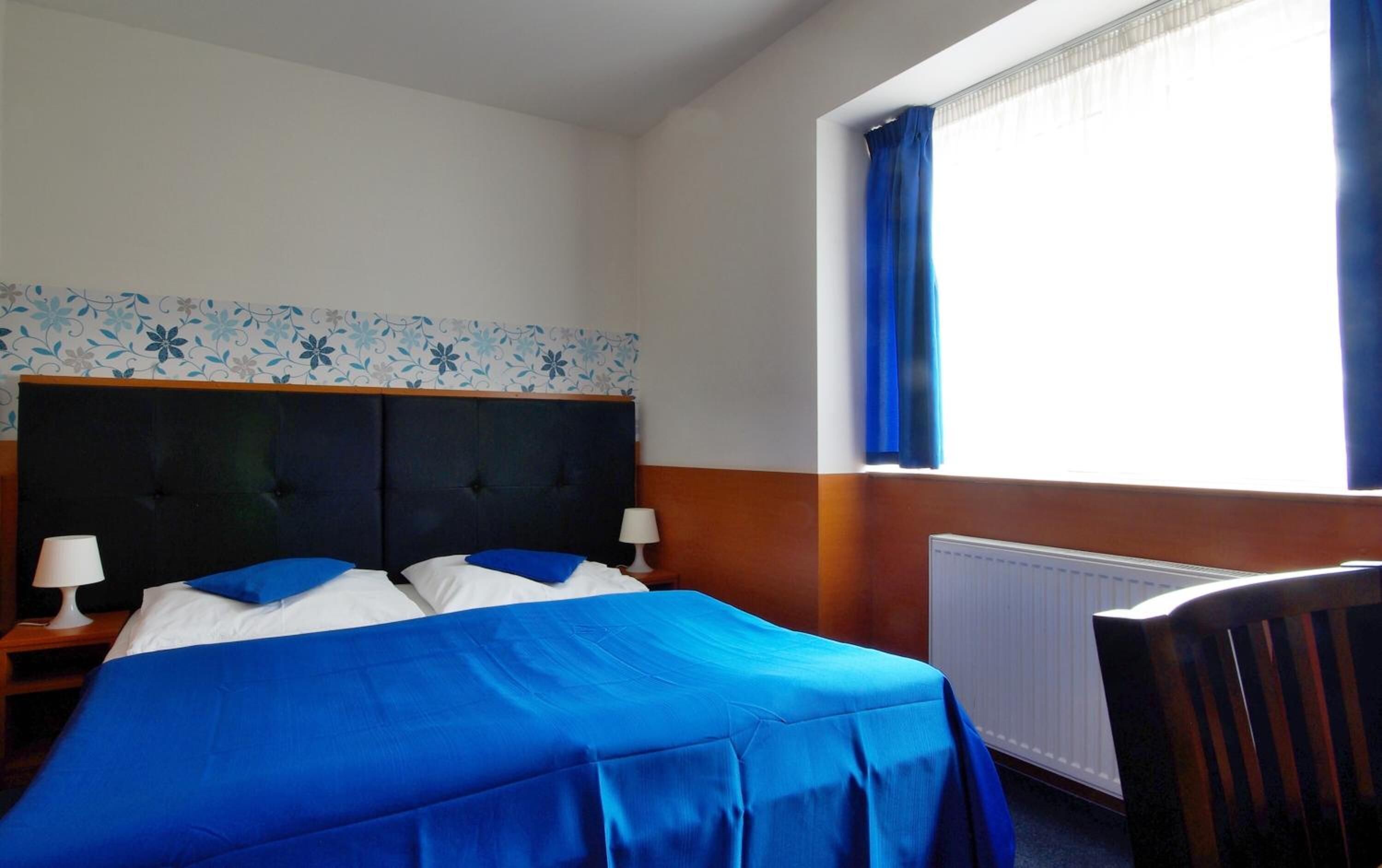 A hotel room with a large bed, two bedside tables, a radiator, and a window with blue curtains.