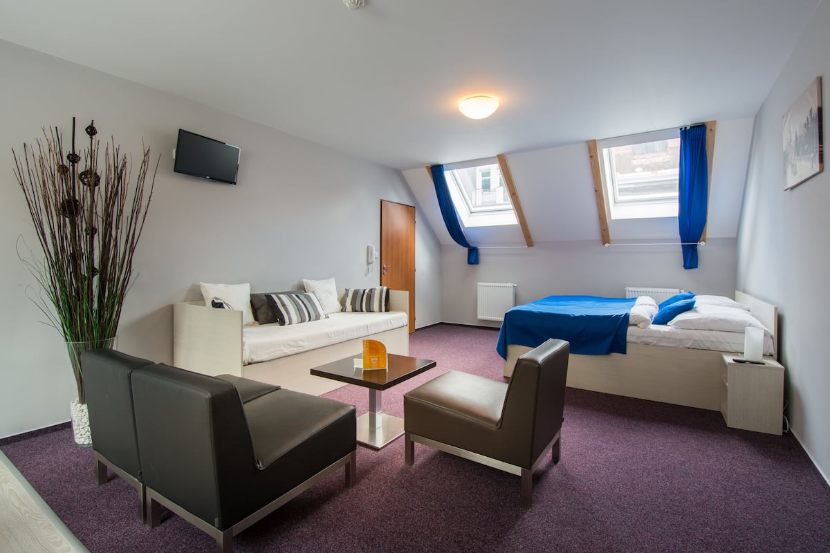Camera d'albergo compatta con divano, un piccolo tavolo, una sedia, un letto con copriletto blu, una TV a parete e una finestra con tende blu.